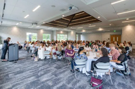 Conference attendees speaking around their table while someone presents at a podium