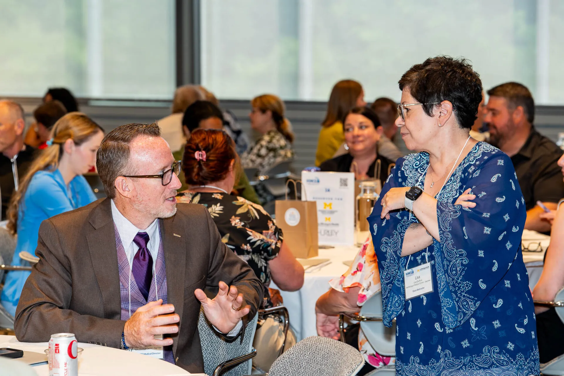 A picture of a woman in blue standing while speaking to a man sitting at a table during a networking session at a conference.