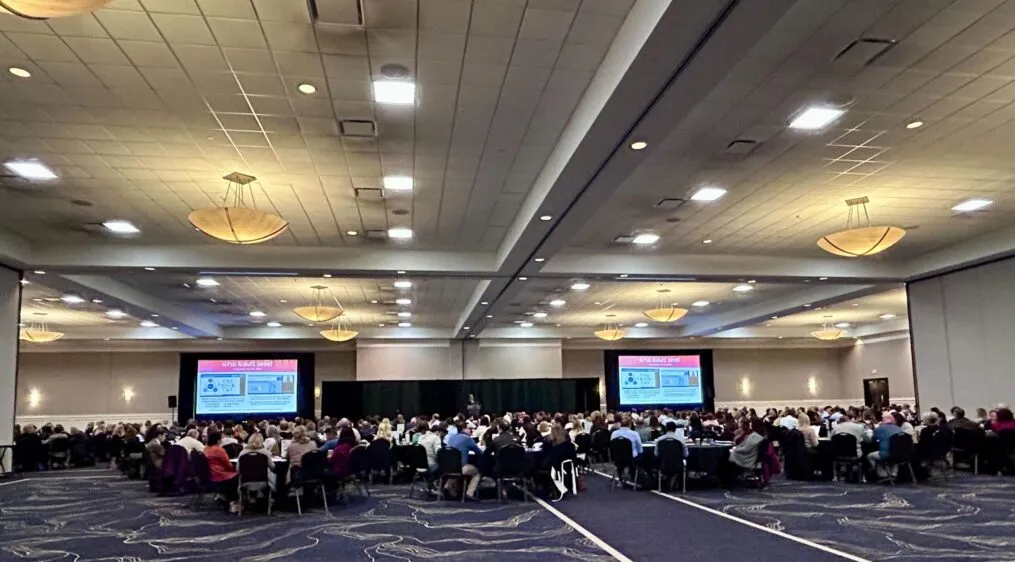 A room with people seated at tables with presentations screens on the left and right