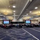 A room with people seated at tables with presentations screens on the left and right