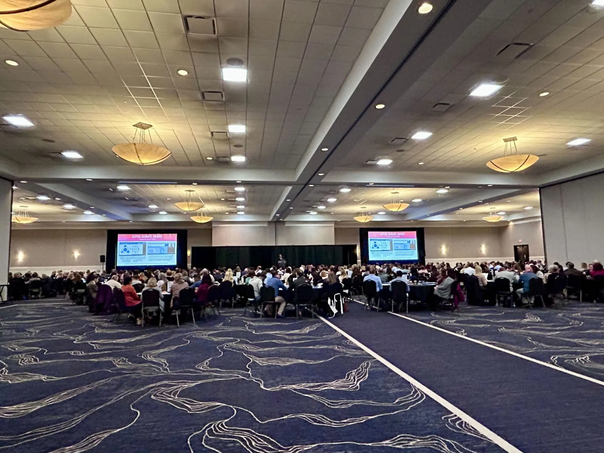 A room with people seated at tables with presentations screens on the left and right