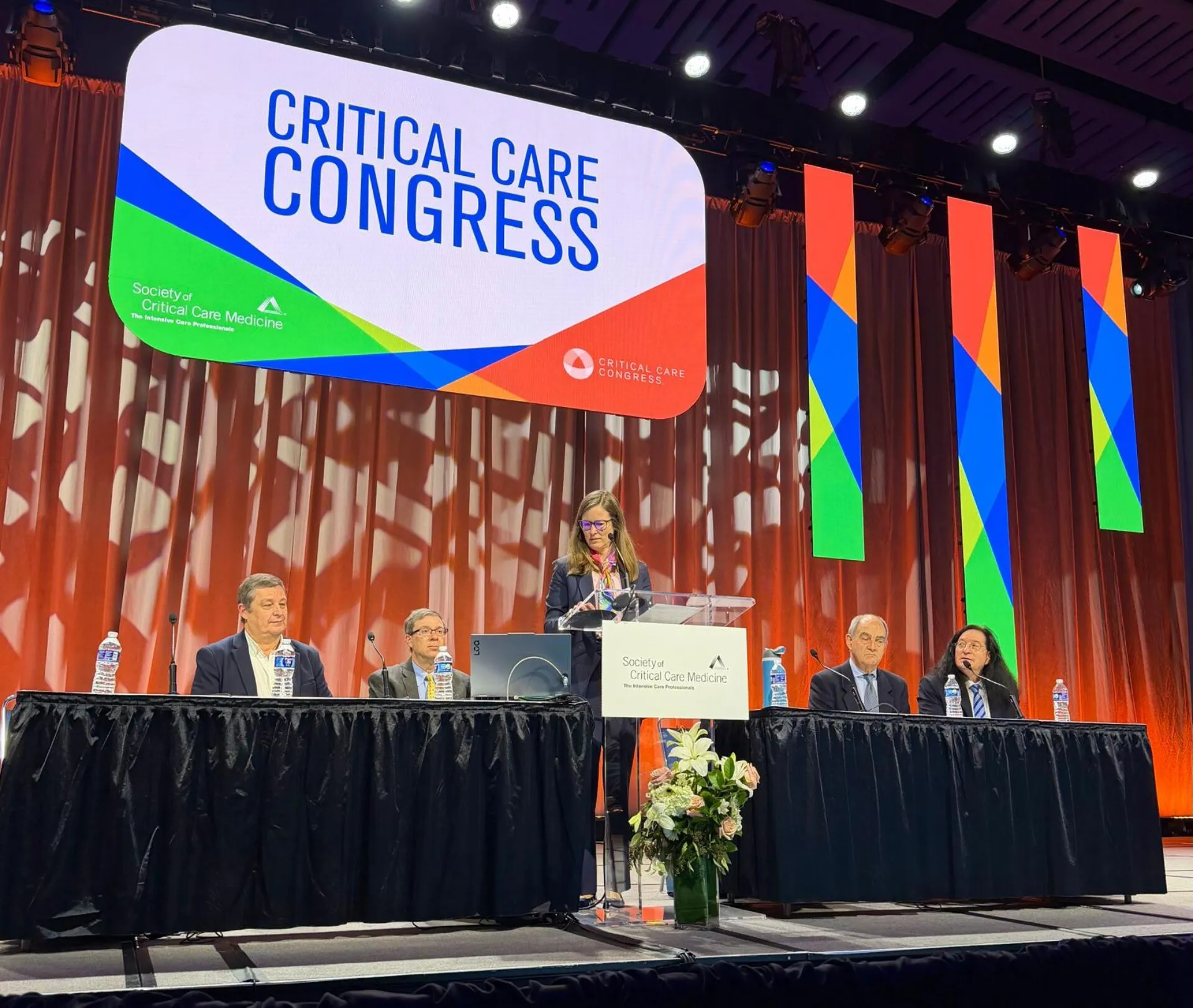 A woman standing in the center of the stage, at a podium, delivering a speech. There are two people seated on both sides of her at a panel table. A large sign hangs above them titled, 
