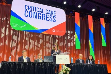 A woman standing in the center of the stage, at a podium, delivering a speech. There are two people seated on both sides of her at a panel table. A large sign hangs above them titled, 