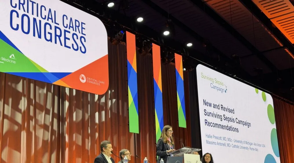 A woman standing in the center of the stage, at a podium, delivering a speech. There are two people seated on both sides of her at a panel table. A large sign hangs above them titled, 