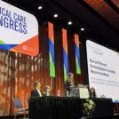 A woman standing in the center of the stage, at a podium, delivering a speech. There are two people seated on both sides of her at a panel table. A large sign hangs above them titled, 