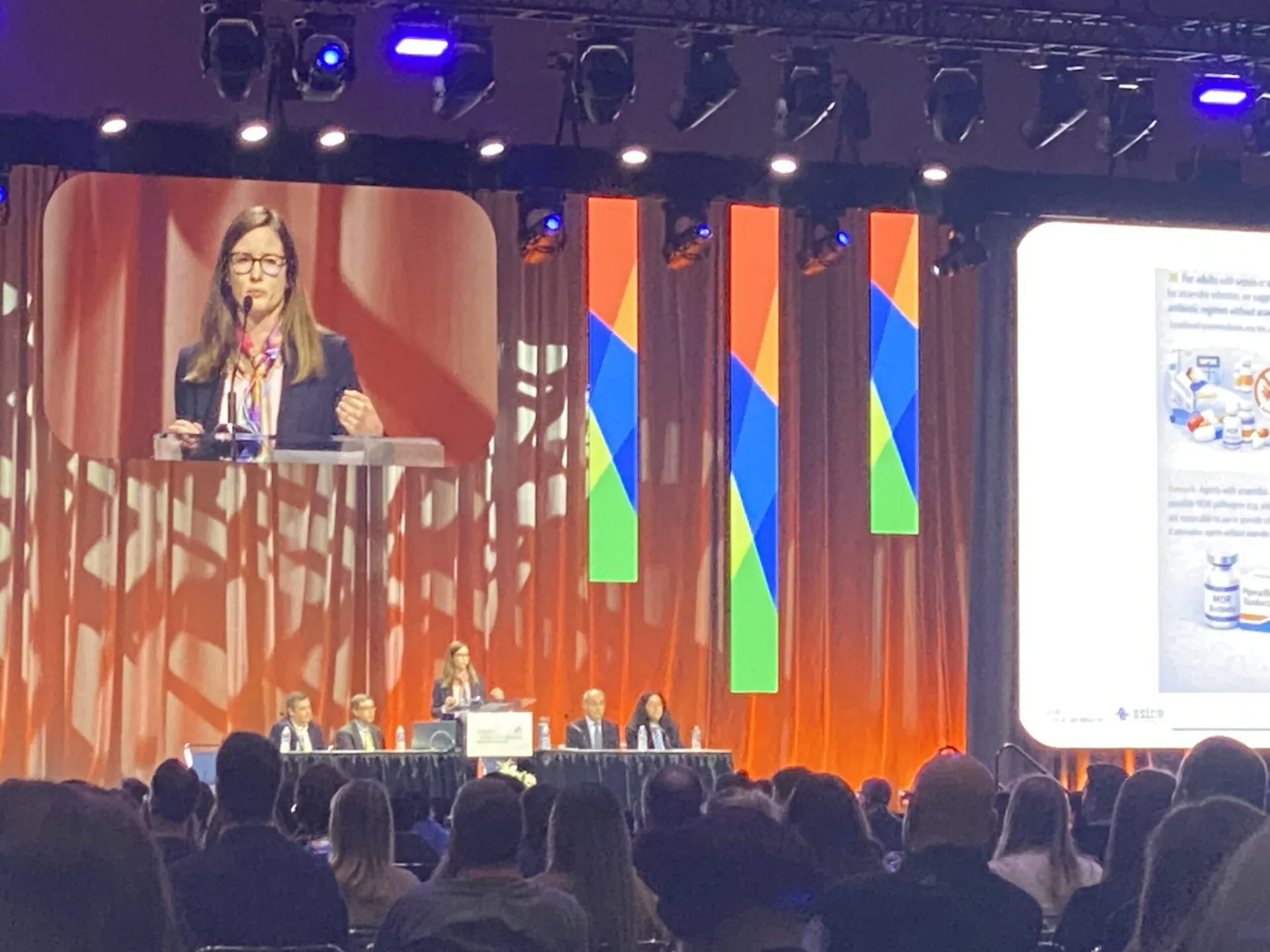 A woman standing in the center of the stage, at a podium, delivering a speech. There are two people seated on both sides of her at a panel table. A large sign hangs above them titled, 
