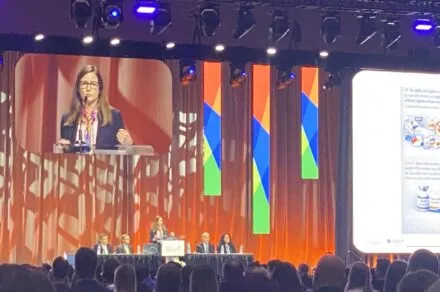 A woman standing in the center of the stage, at a podium, delivering a speech. There are two people seated on both sides of her at a panel table. A large sign hangs above them titled, 
