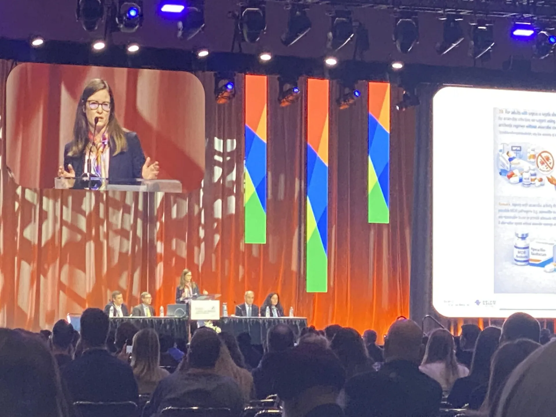 A woman standing in the center of the stage, at a podium, delivering a speech. There are two people seated on both sides of her at a panel table. A large sign hangs above them titled, 