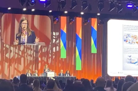 A woman standing in the center of the stage, at a podium, delivering a speech. There are two people seated on both sides of her at a panel table. A large sign hangs above them titled, 