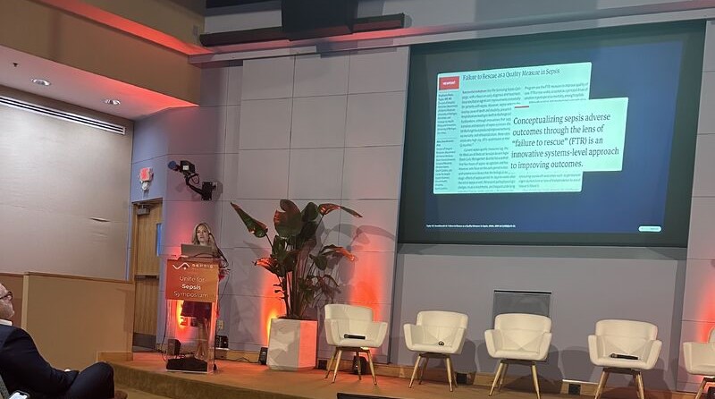 Dr. Parks Taylor speaks at a podium at the Unite for Sepsis Symposium. Behind her, a projected slide displays text reading "Conceptualizing sepsis adverse outcomes through the lens of 'failure to rescue' (FTR) is an innovative systems-level approach to improving outcomes." Empty white panel chairs are arranged on stage.